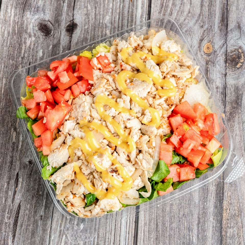 Grilled chicken salad with lettuce, diced tomatoes, onions, and yellow dressing in a clear container on a rustic wooden table.