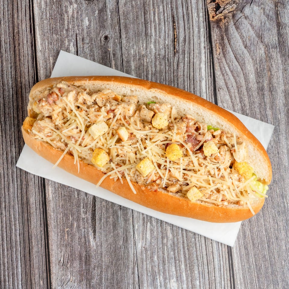 Toasted sub sandwich filled with creamy chicken, bacon, lettuce, shredded cheese, and croutons on a wooden table.