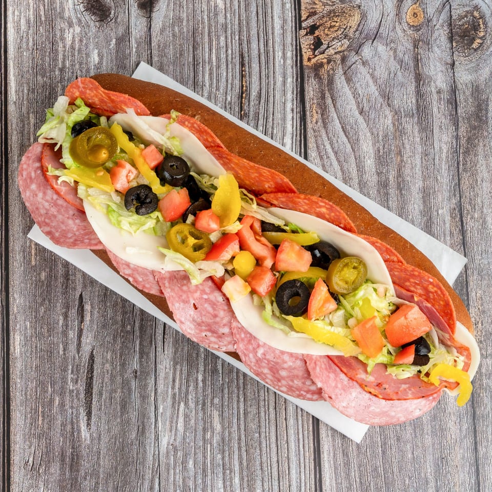 Italian sub sandwich filled with salami, pepperoni, cheese, lettuce, tomatoes, olives, and jalapeños on a rustic wooden table.