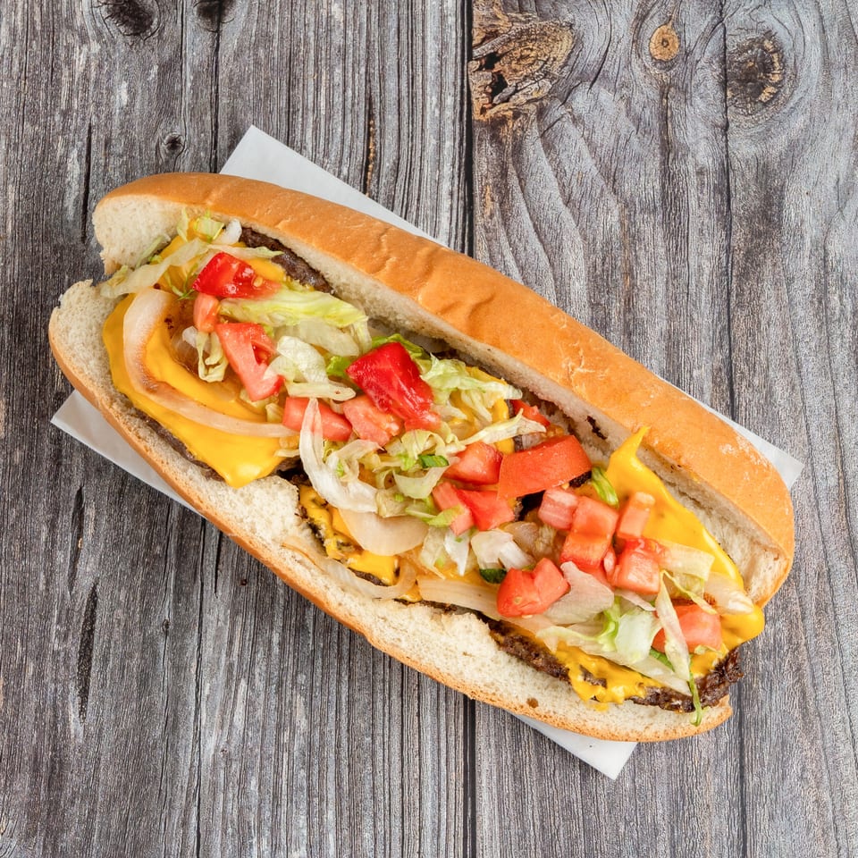Cheeseburger sandwich on a long toasted bun, topped with lettuce, tomatoes, onions, and melted cheese on a rustic wooden table.