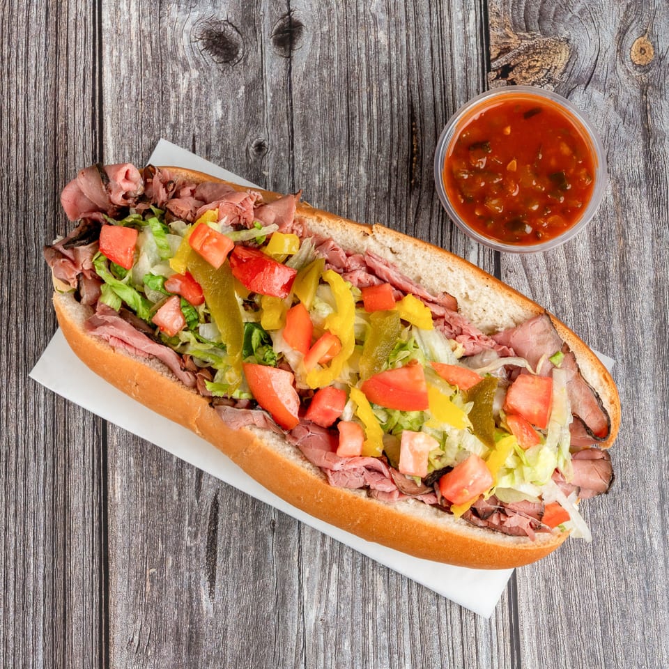 Overhead view of a roast beef sub topped with lettuce, tomatoes, peppers, and a cup of red salsa on a rustic wooden table.