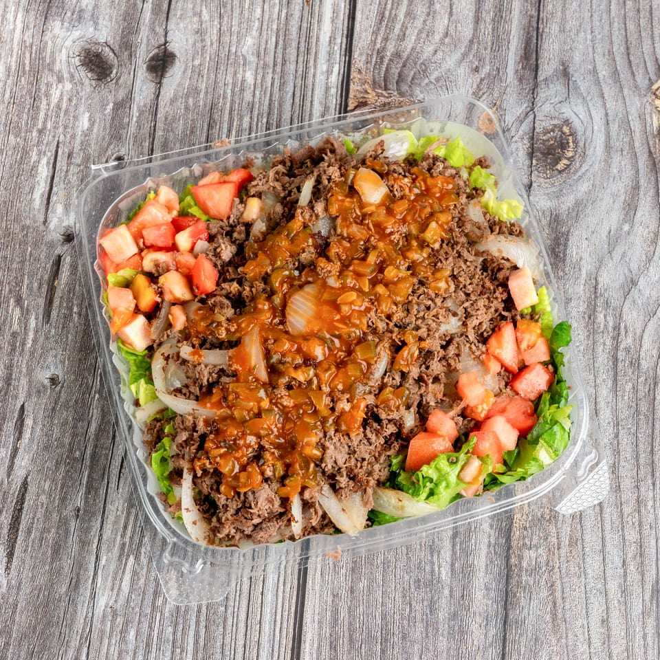 Takeout container filled with lettuce, seasoned ground beef, grilled onions, diced tomatoes, and reddish sauce on a rustic wooden table.