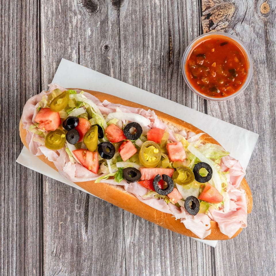 Submarine sandwich topped with ham, lettuce, tomatoes, black olives, onions, pickles, and jalapeños beside a cup of red sauce.